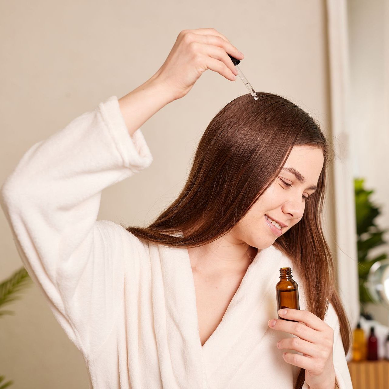 A woman in a bathrobe is applying a serum to her hair.