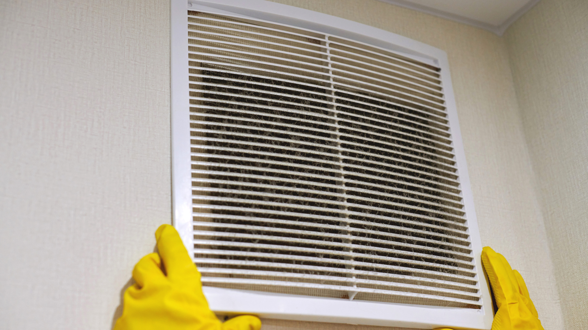 A person wearing yellow gloves is cleaning a vent on a wall.