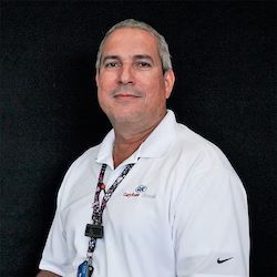 Angel Melendez wearing a white shirt and a lanyard is standing in front of a black background.