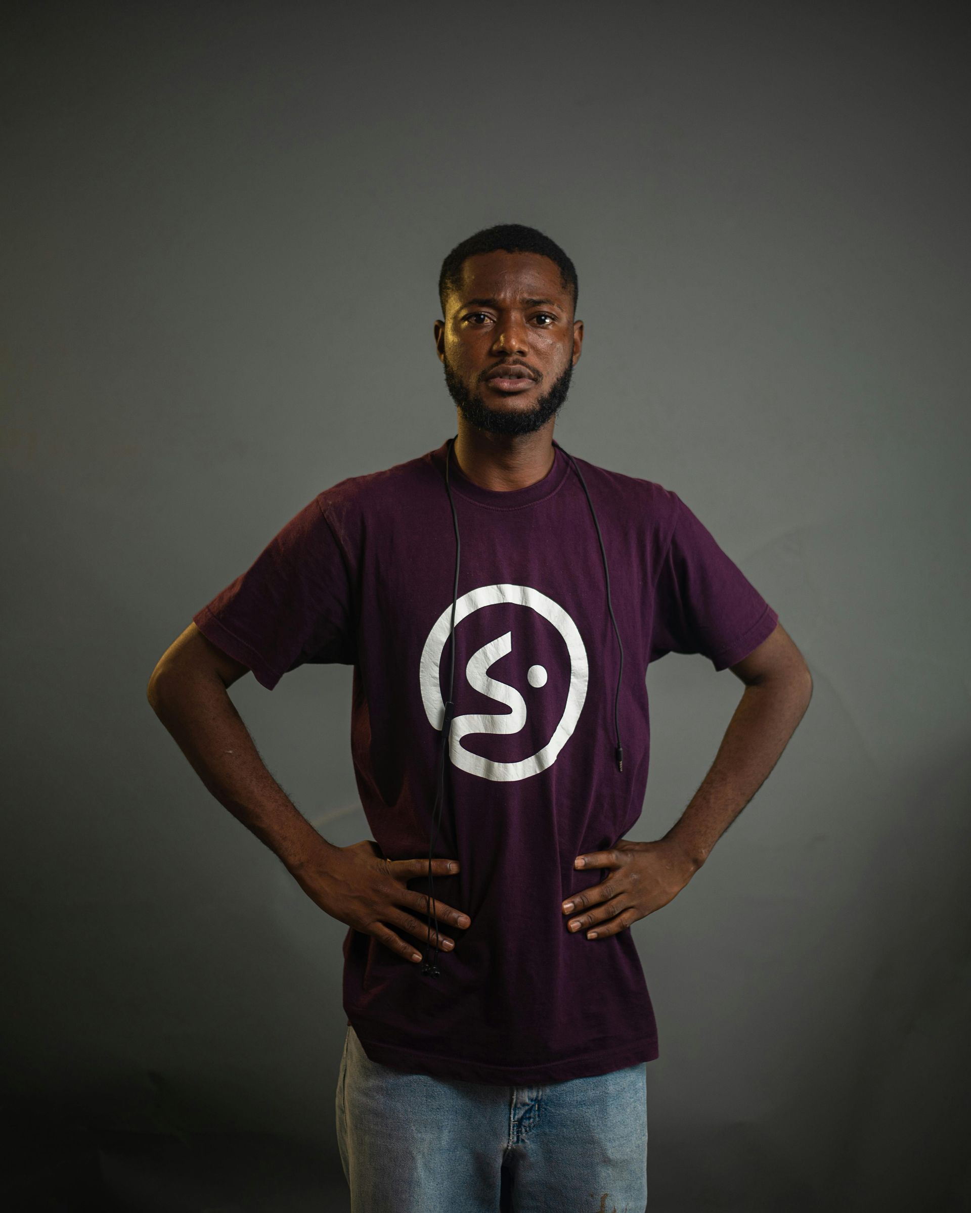 Man in purple shirt with logo, hands on hips, looking surprised, gray background.