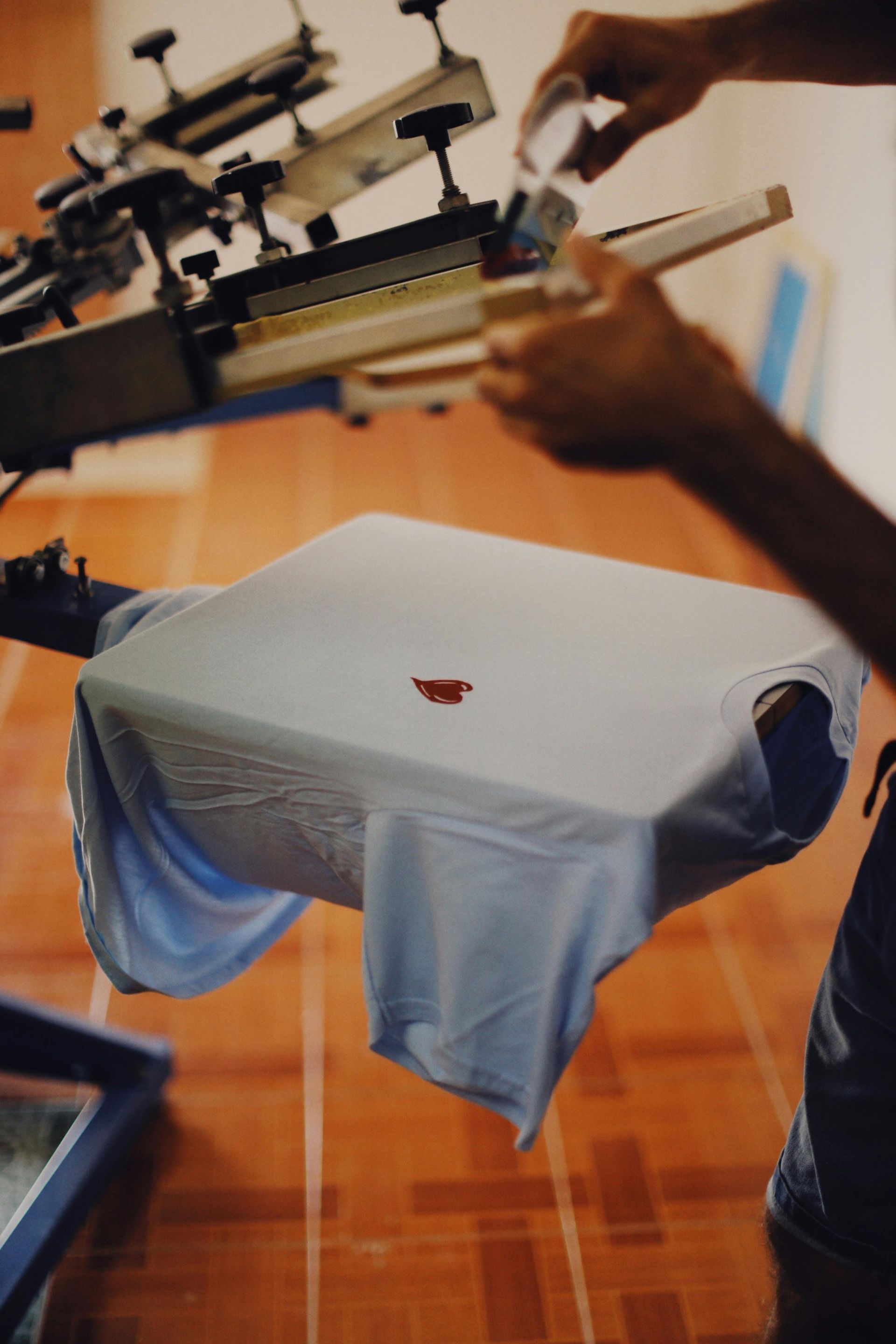 A person screen printing a red design onto a light blue t-shirt with a printing machine in a tiled room.