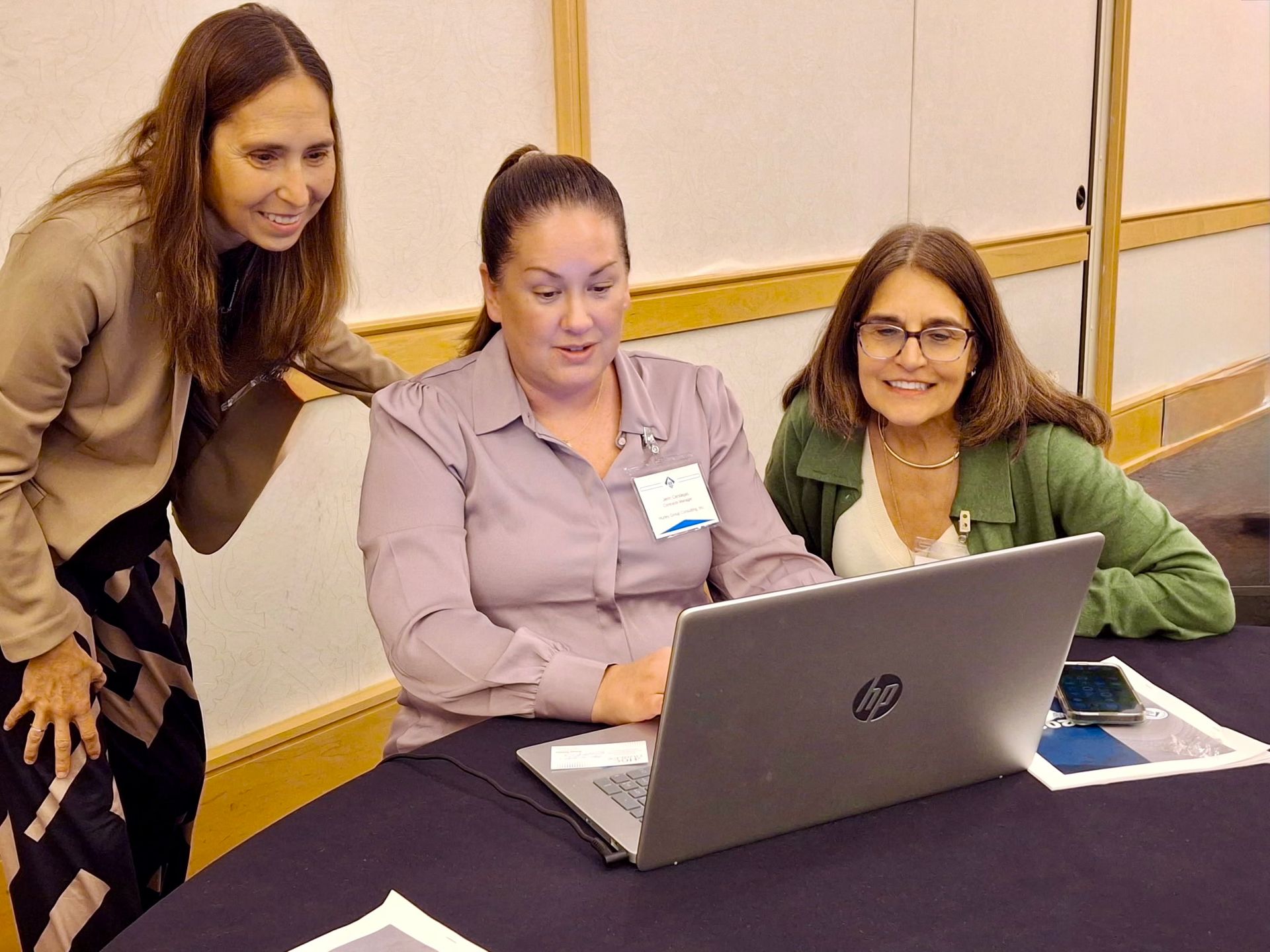 Three women looking at a laptop, one points, others smile, in a room with a blue tablecloth.