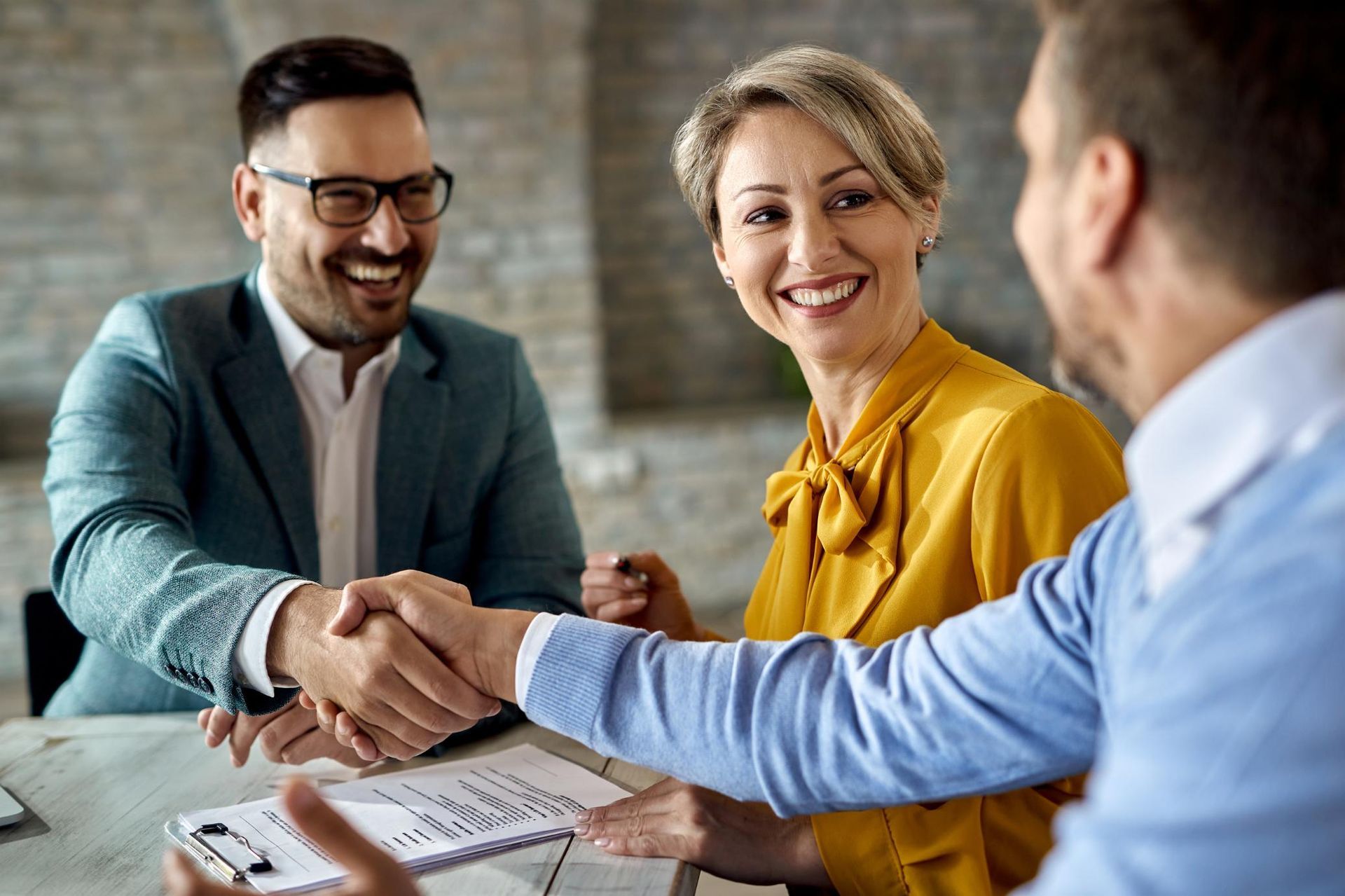 Three people at a table; two shaking hands. Man in glasses smiles, woman in yellow smiles.