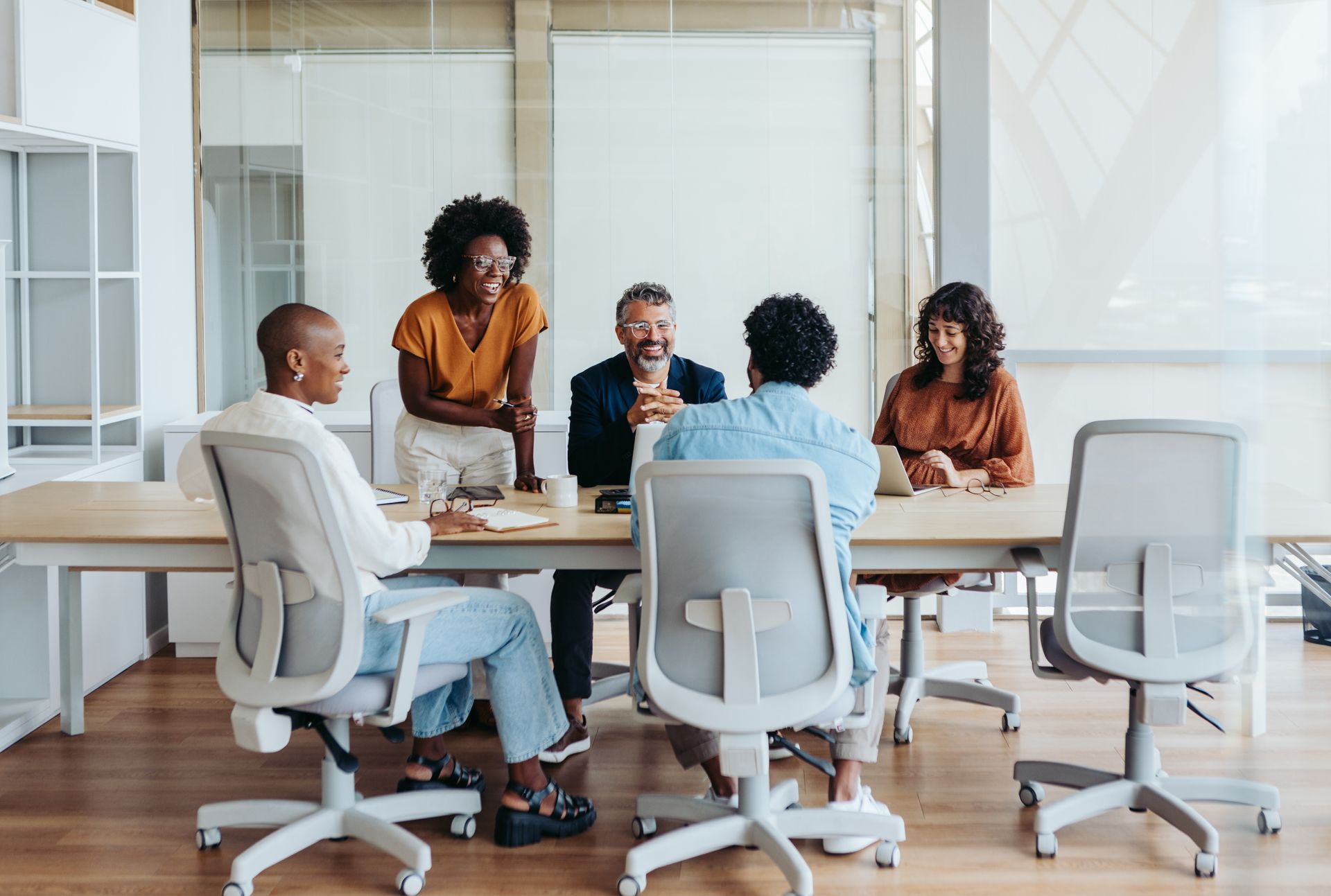 Group of people at a conference table, some looking at laptops, others listening. 