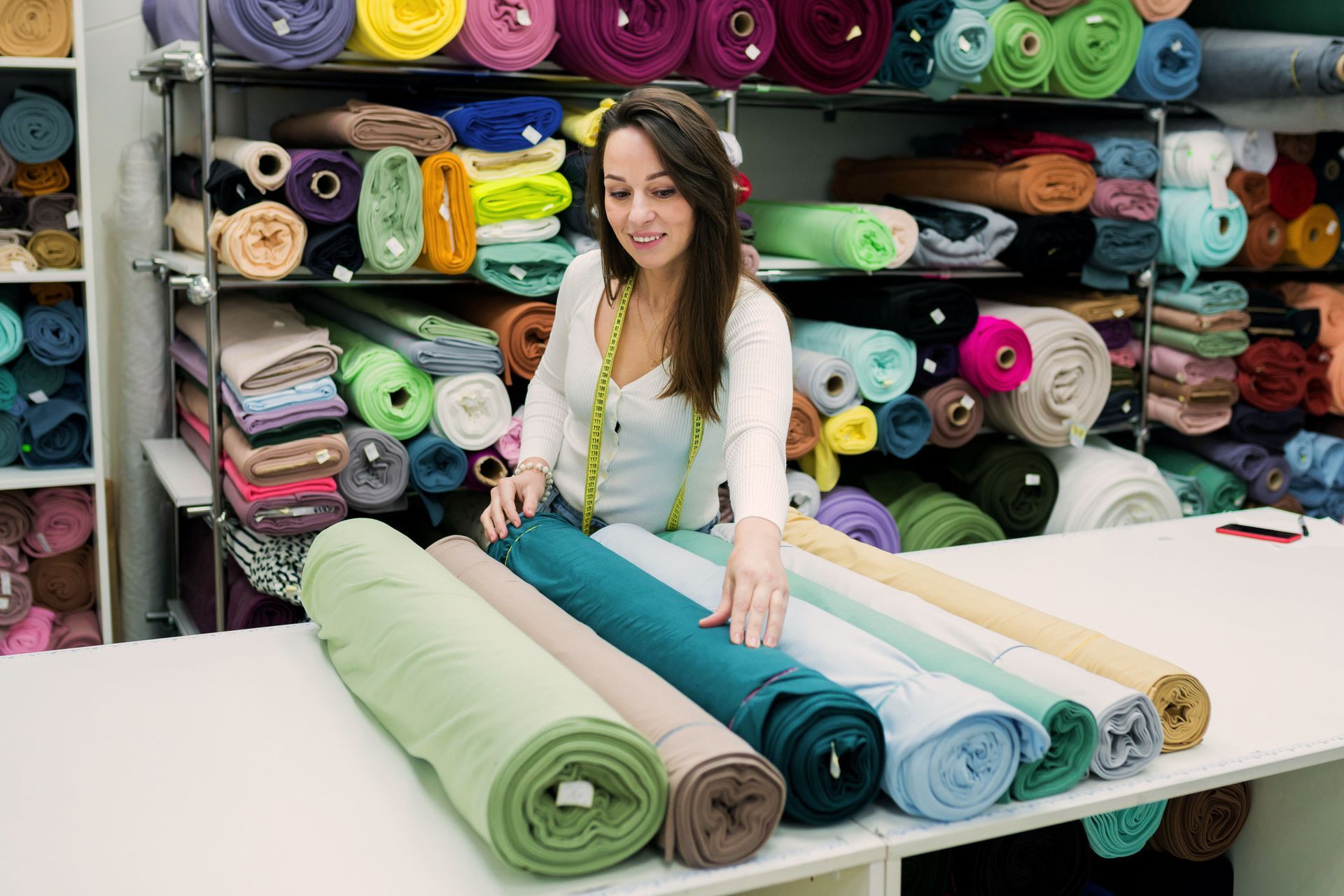 Woman arranging fabric rolls at a shop counter, surrounded by shelves of colorful textiles.