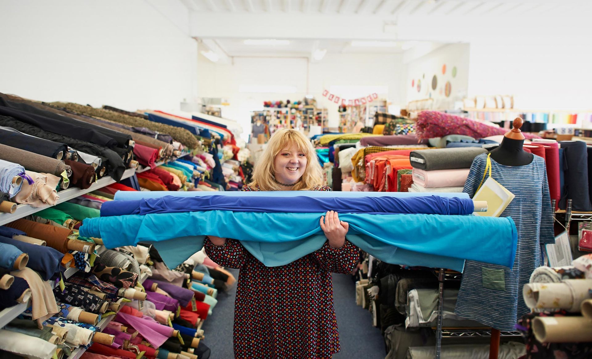 Woman holding blue fabric rolls in a fabric store, smiling. Many fabric bolts on shelves.