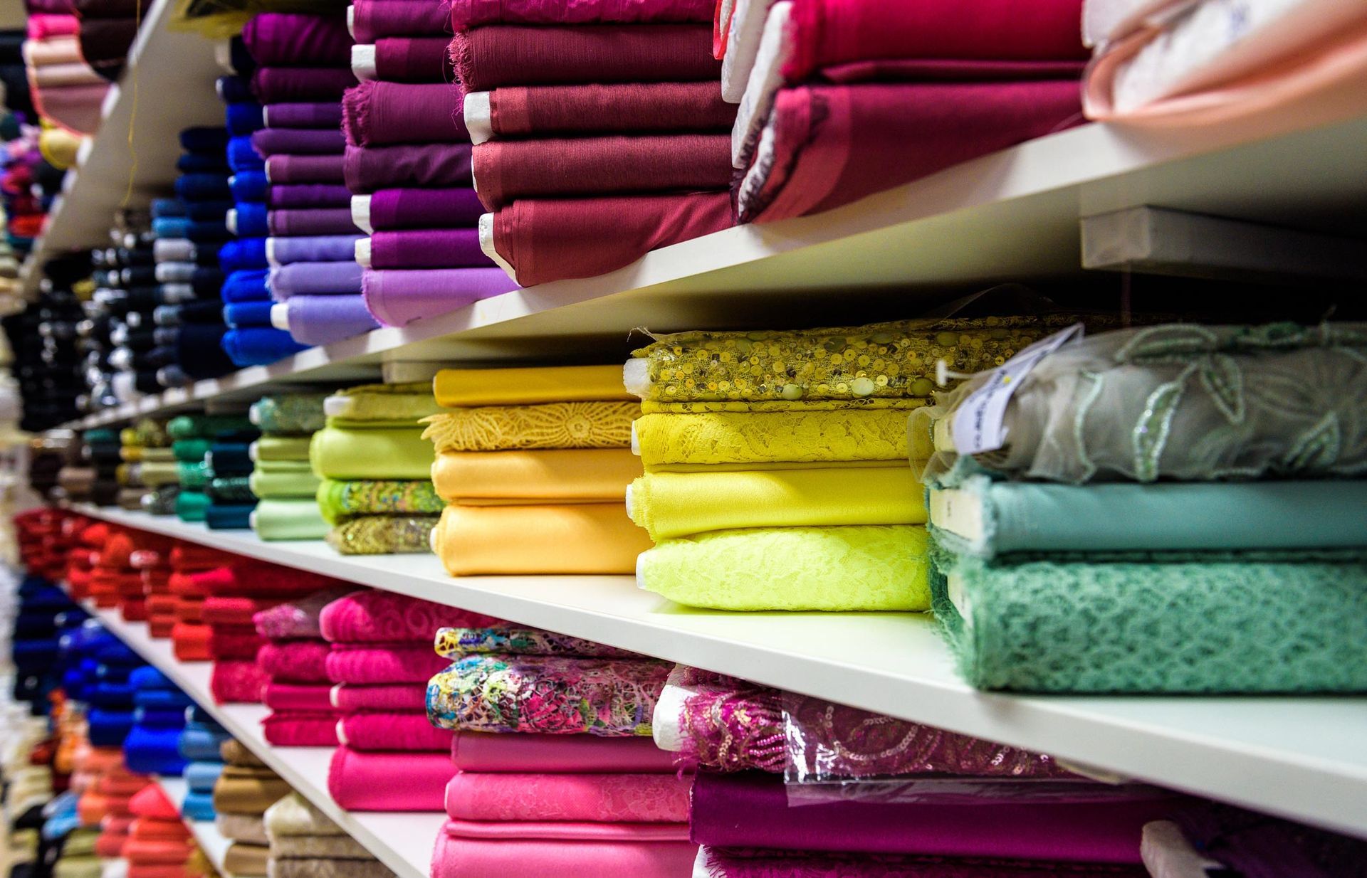 Shelves stacked with colorful fabric bolts in a store. Reds, pinks, blues, greens, yellows, and purples are visible.