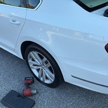 White car with a flat tire; a wrench and tool box lay beside it on pavement.
