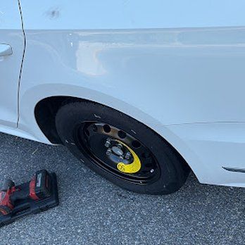 White car with a flat tire replaced by a black spare tire; an electric wrench is on the ground.