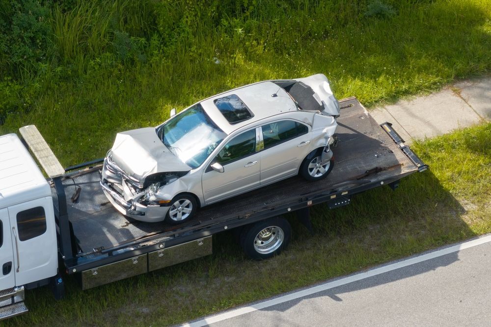 Damaged silver car on a flatbed tow truck next to a grassy roadside.