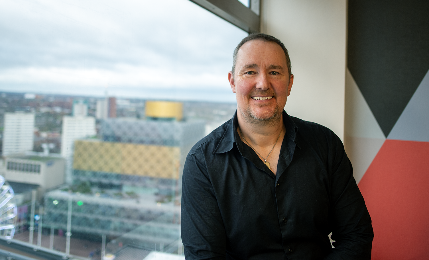 Man smiling, seated by a window overlooking a city. He wears a black shirt, buildings visible outside.