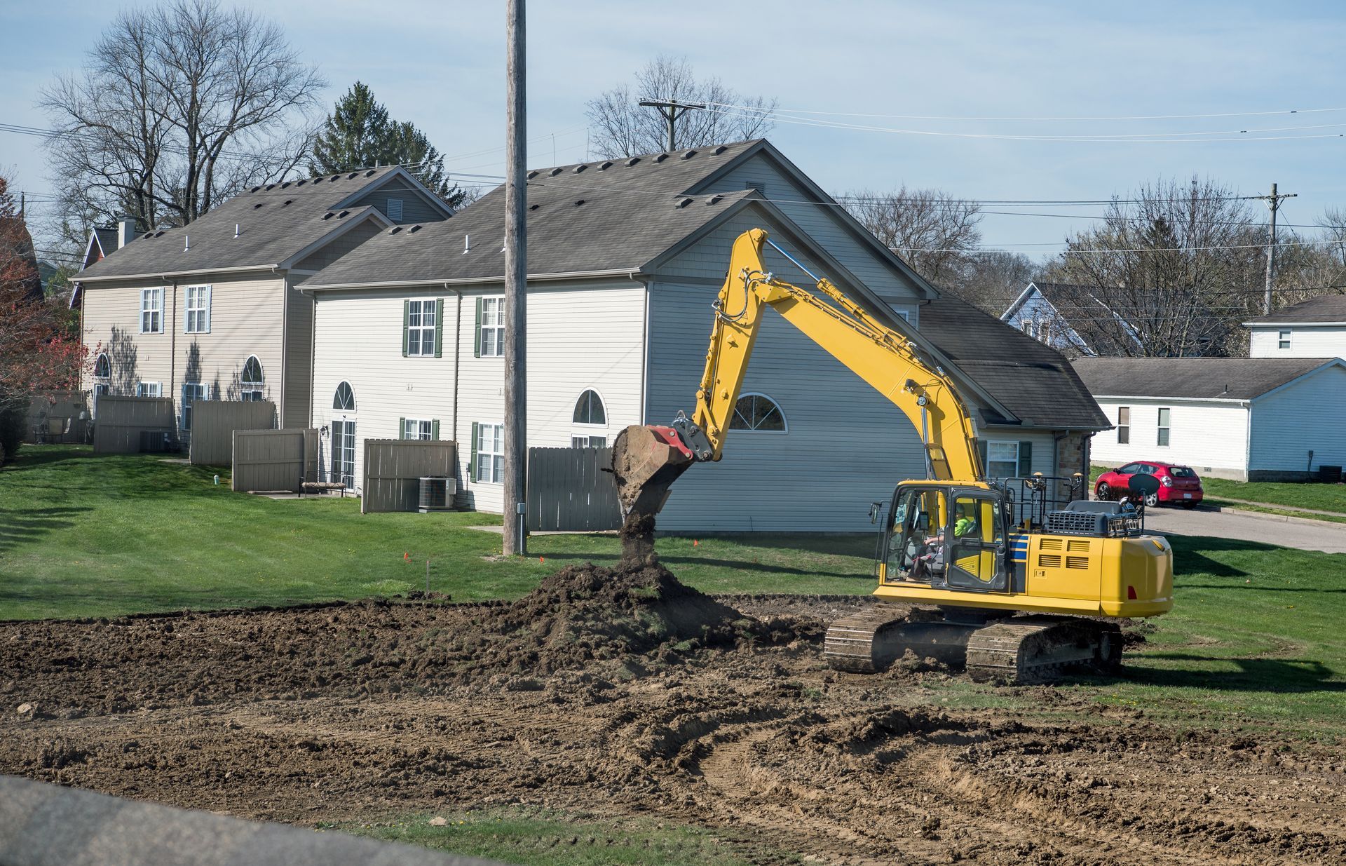 Yellow excavator digging dirt in a suburban yard near white houses.