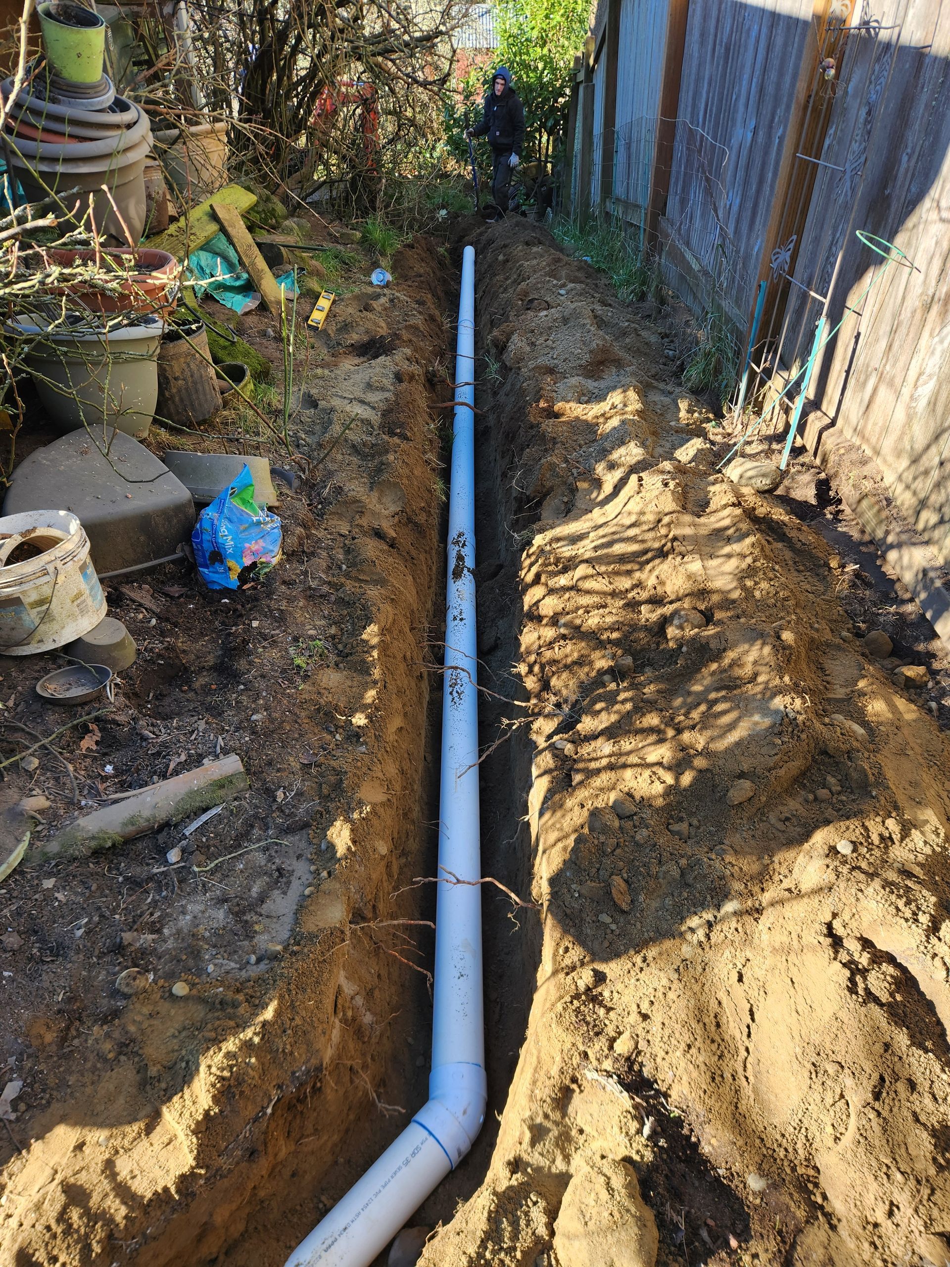 A long trench in a yard with a grey pipe running through it, person in background.