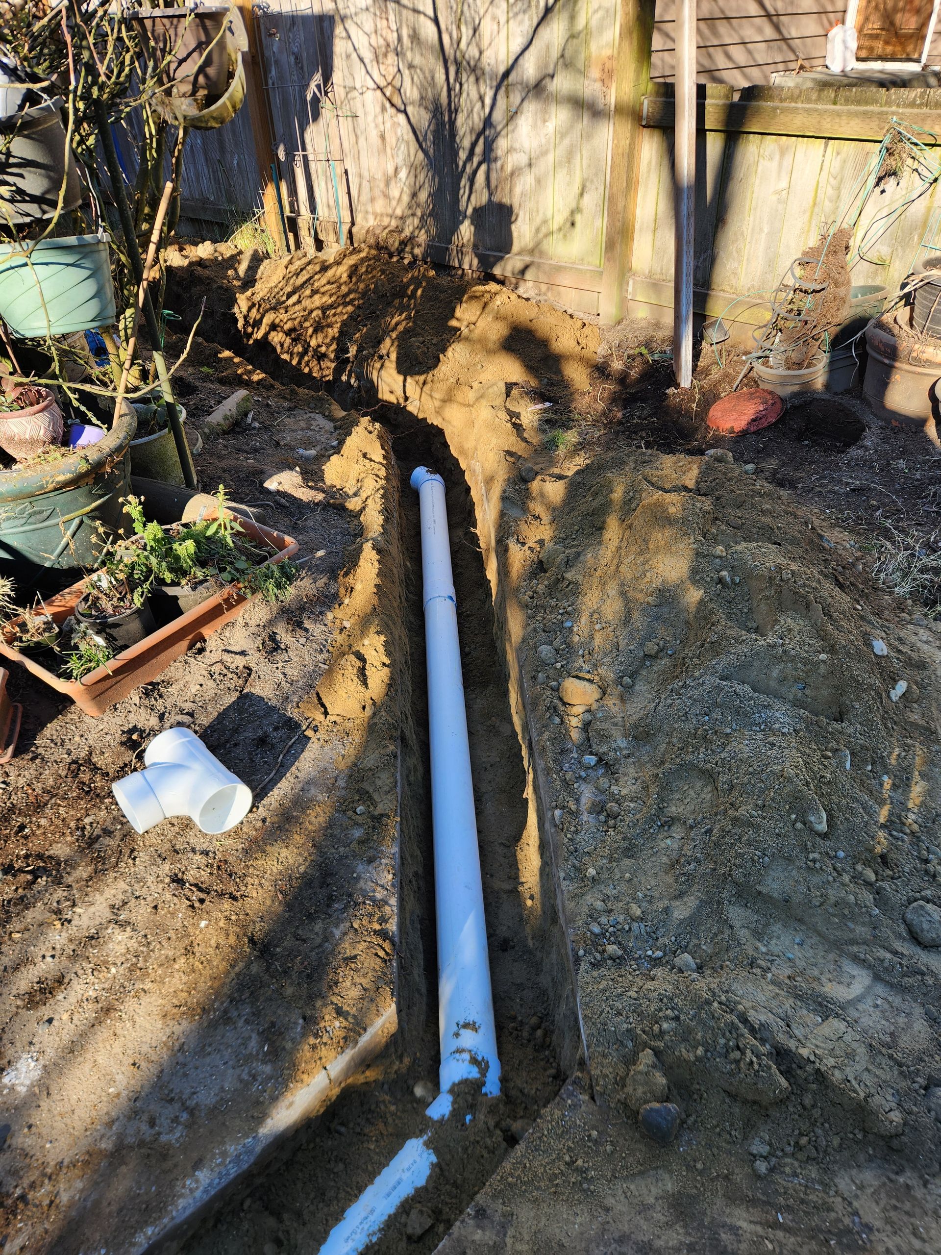 A white PVC pipe lies in a trench dug in dirt, alongside PVC fittings.
