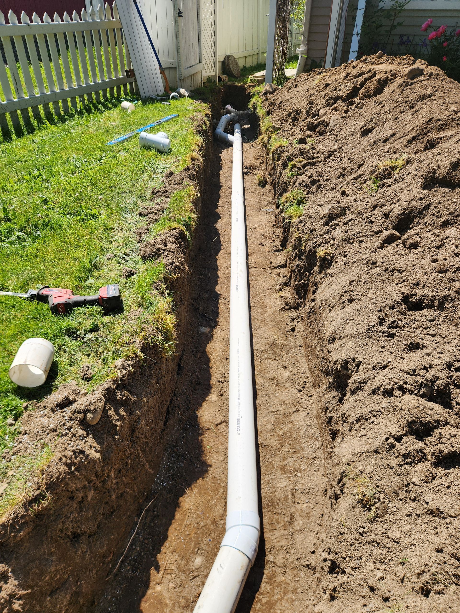 Trench in a yard, holding white PVC pipe. A fence and house are in the background, with tools and grass visible.