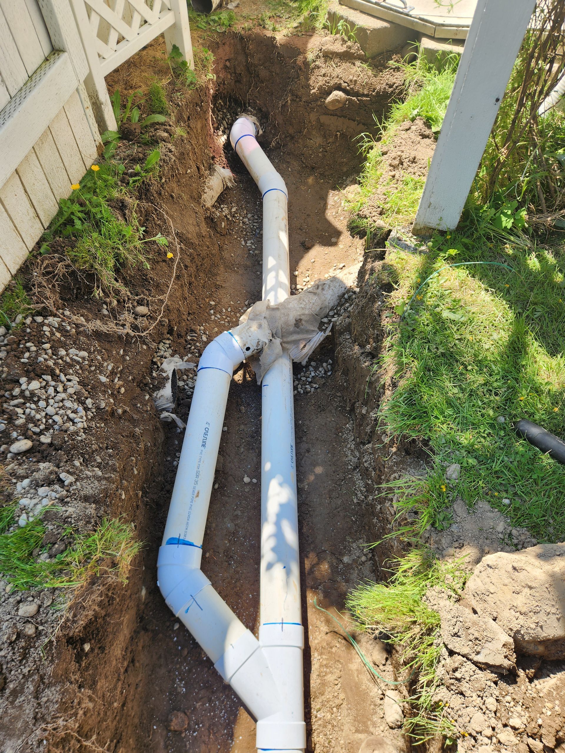 White PVC pipes in a trench, connected and extending upward, near a white fence and grass.