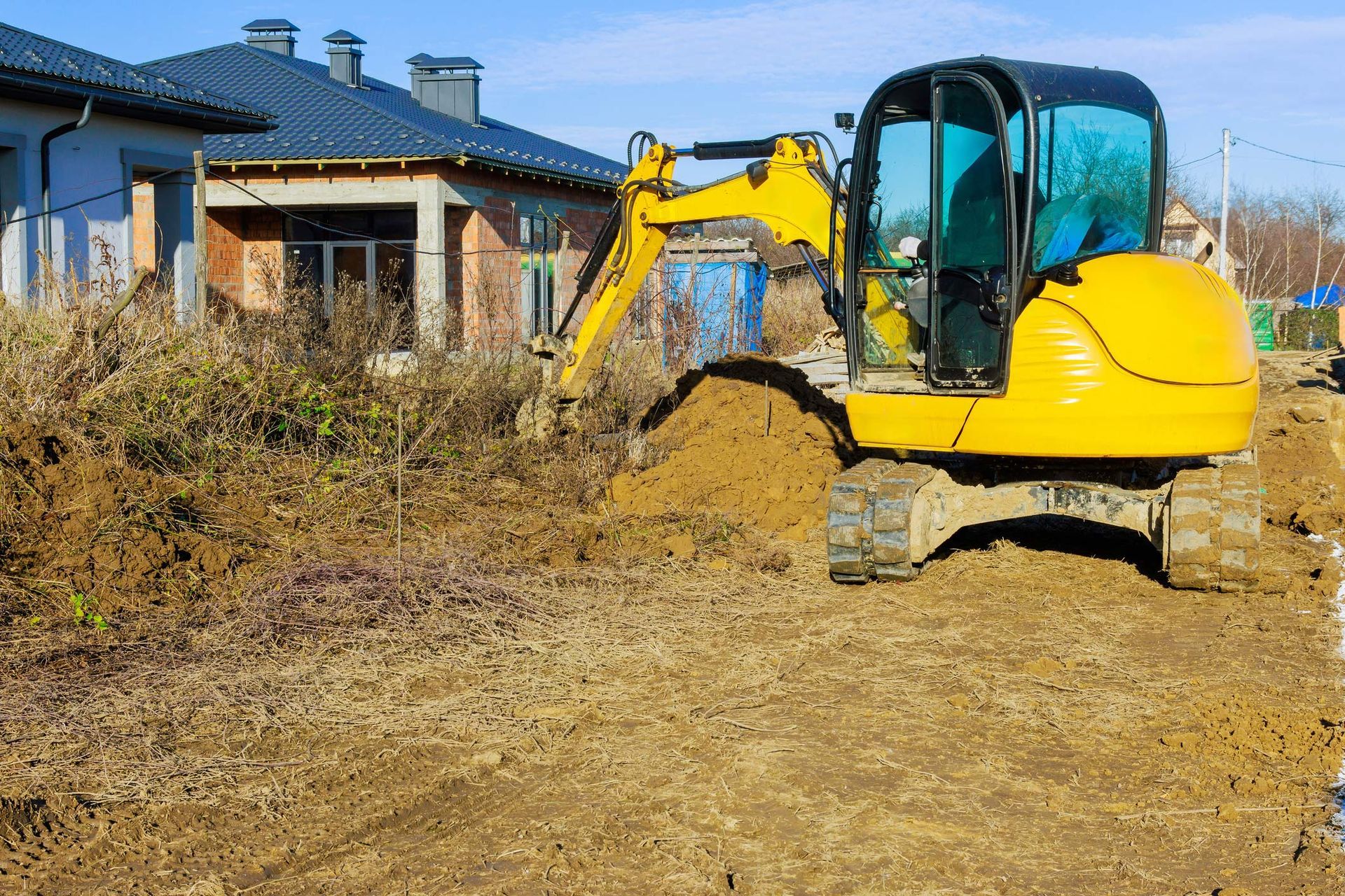 Yellow excavator digging in dirt near a house on a sunny day.