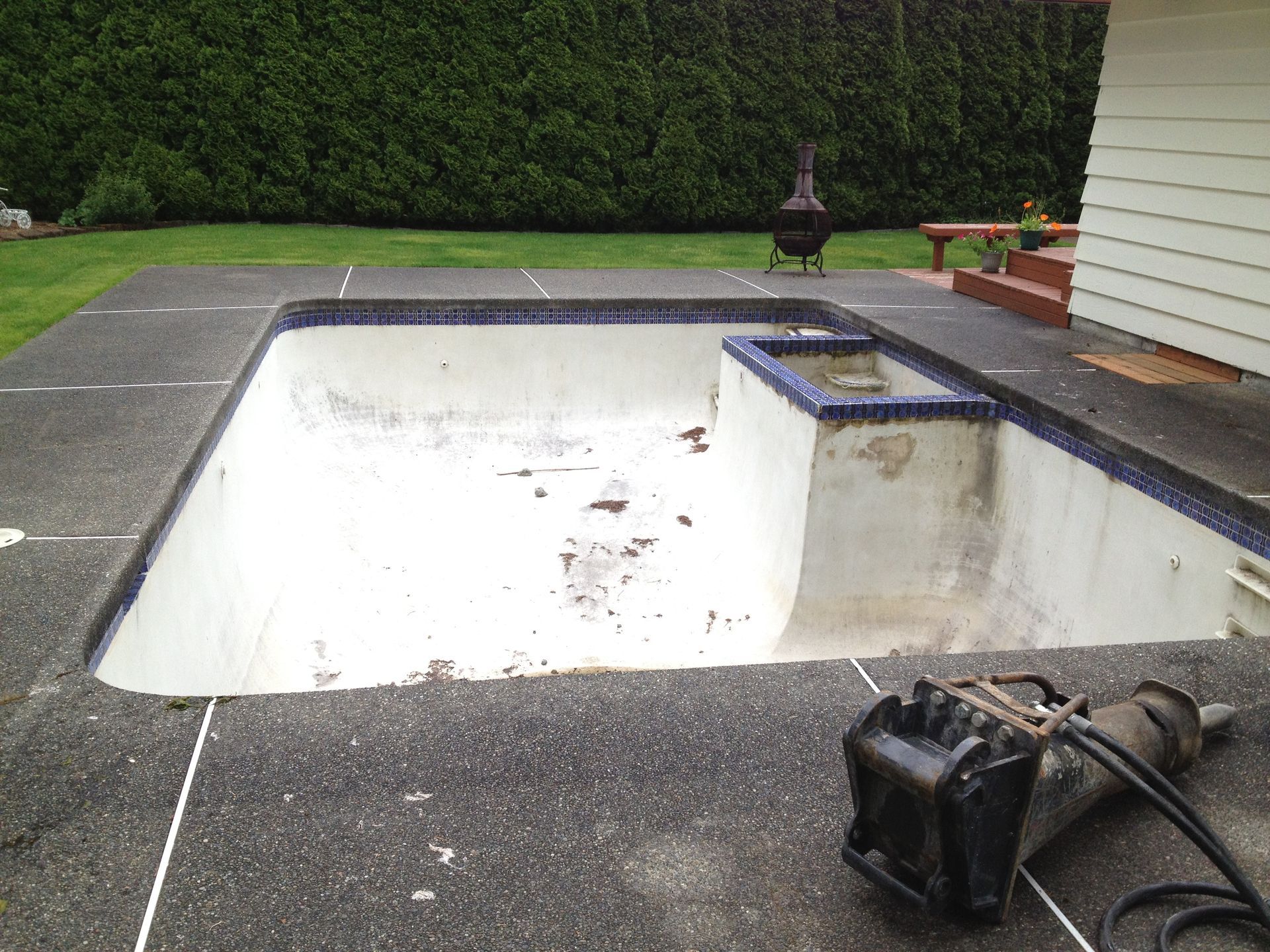 Empty pool with chipped interior, surrounded by concrete patio. A jackhammer rests on the patio.