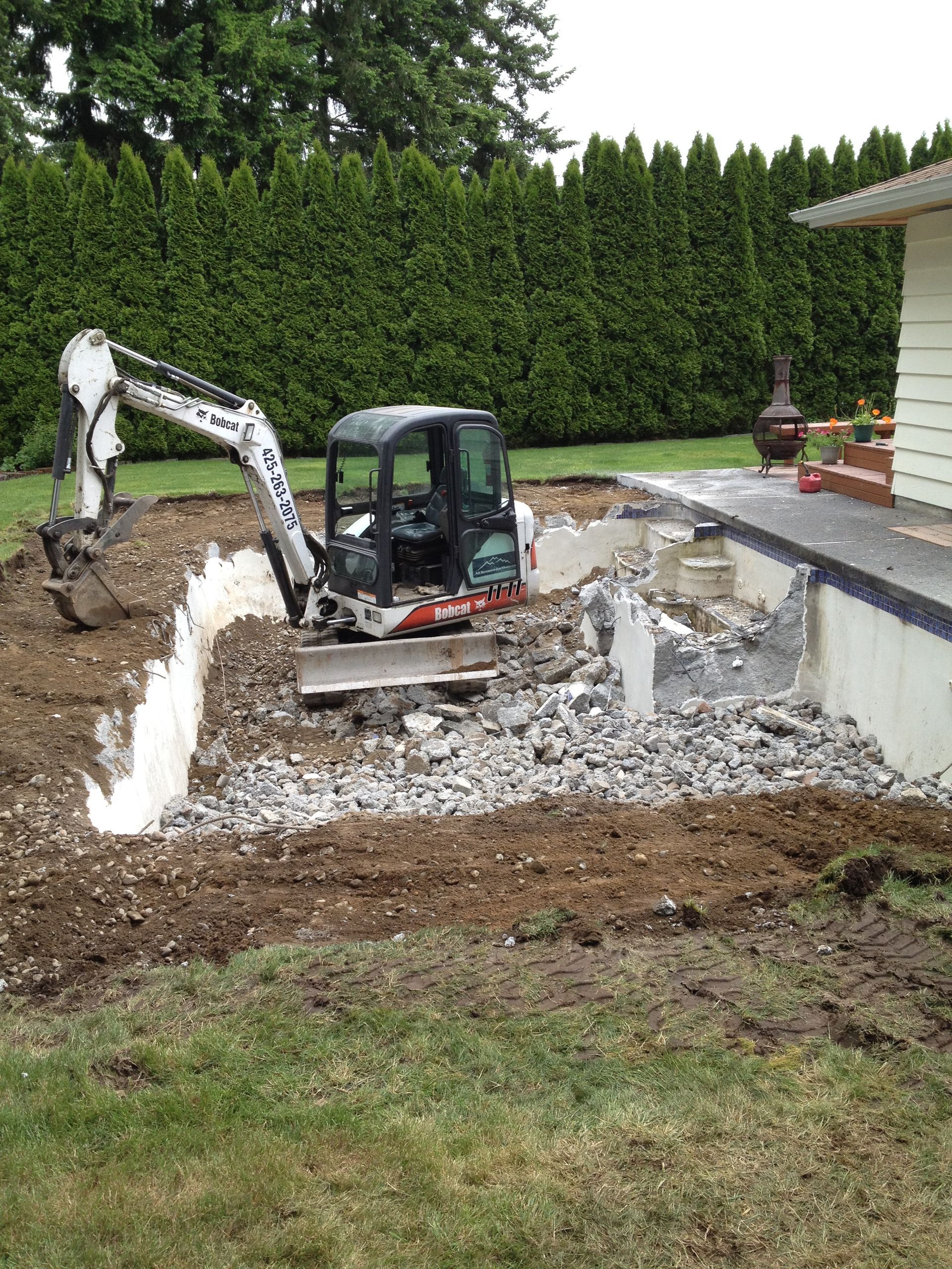 Mini excavator demolishing a concrete structure in a backyard. Earth and rubble surround it, with green lawn and hedge in the background.