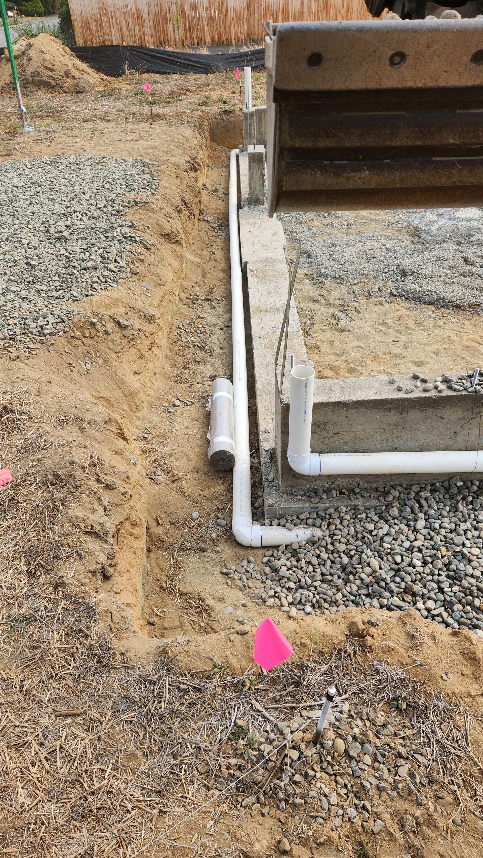Construction site with buried pipes, gravel, and concrete foundation. Pink marker flag.