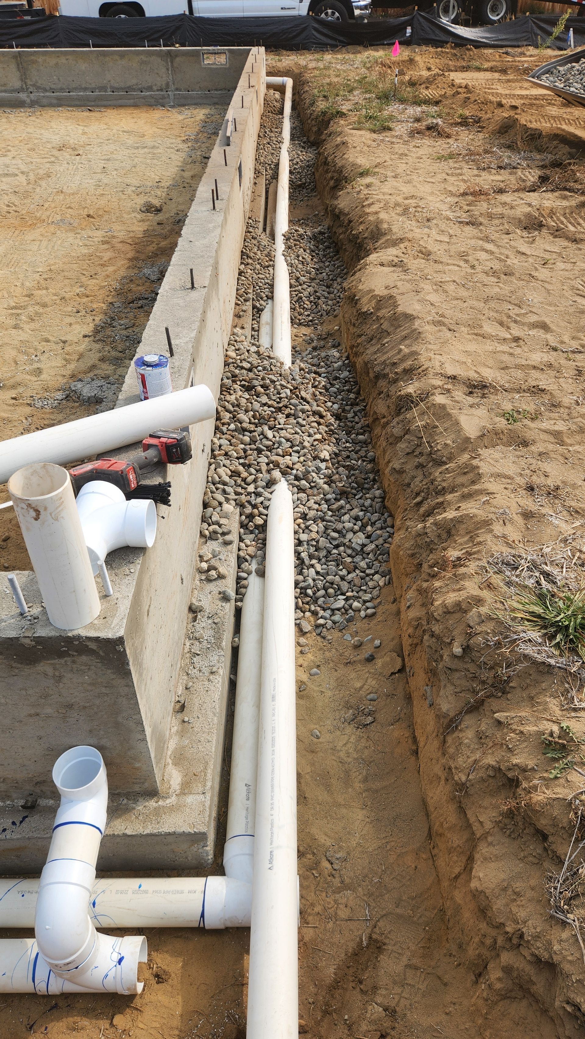 Construction site with buried white PVC pipes, gravel, and a concrete foundation.