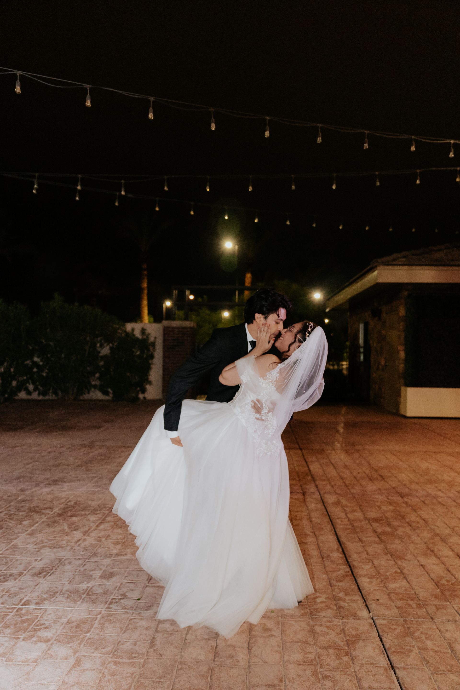 Bride and groom sharing a romantic kiss during their wedding dance at night under string lights.