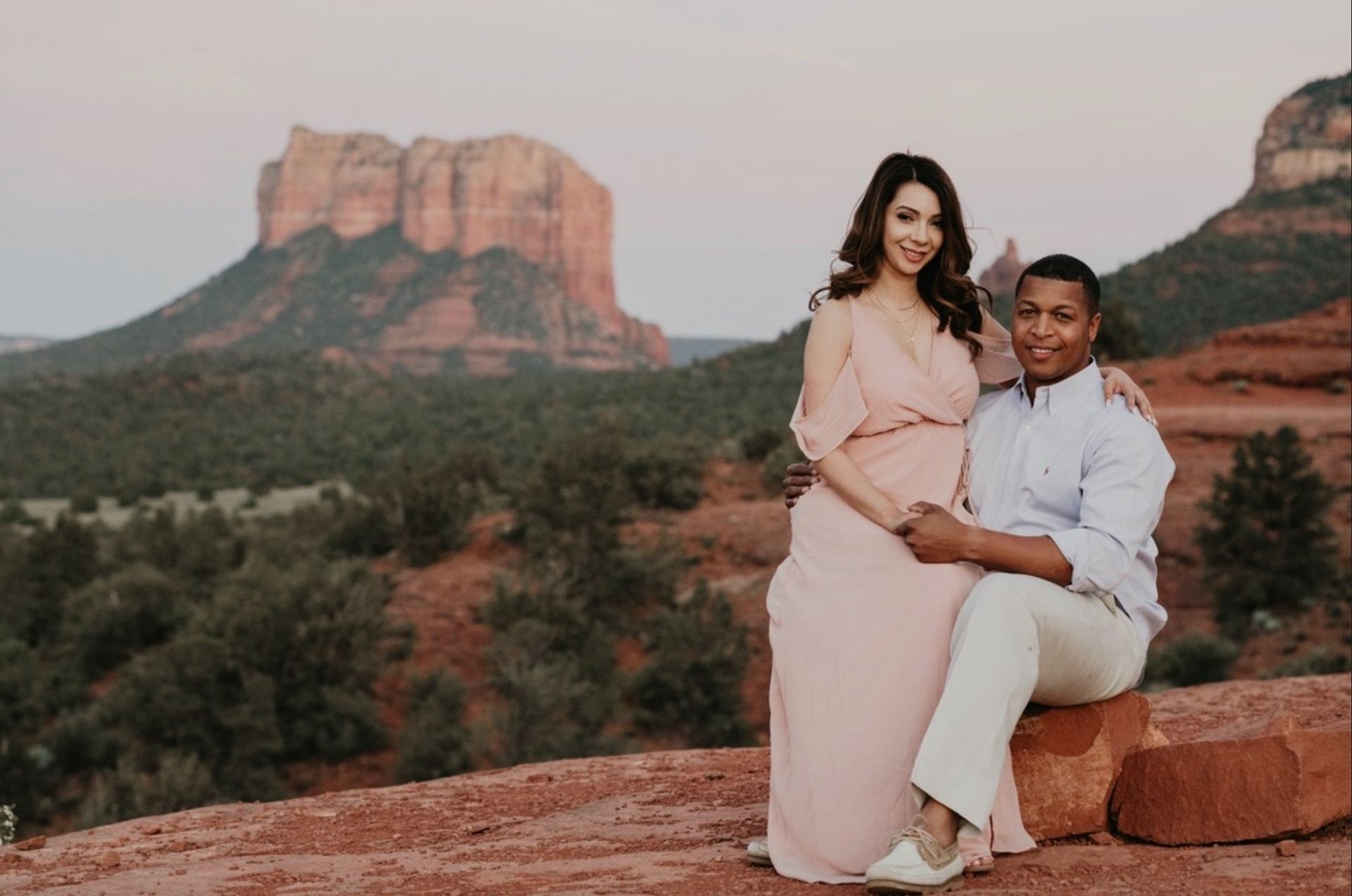 A man and a pregnant woman are sitting on a rock in front of a mountain.