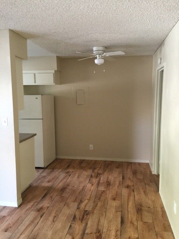 Empty beige room with wood-look floor and ceiling fan, opening into a small kitchen area