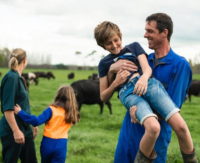 A man is carrying a child in his arms in a field with cows in the background.