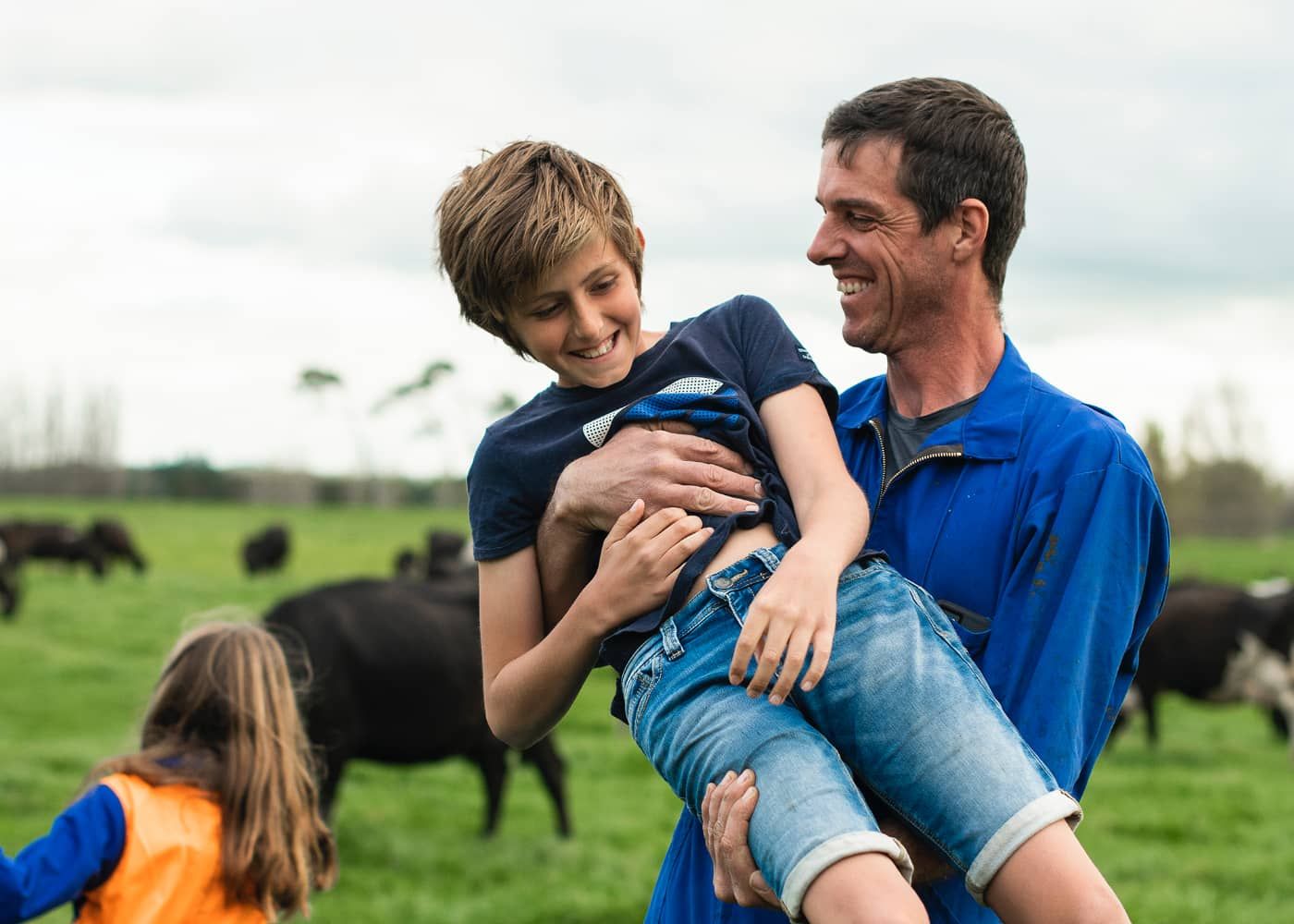 A man is holding a boy in his arms in a field with cows in the background.