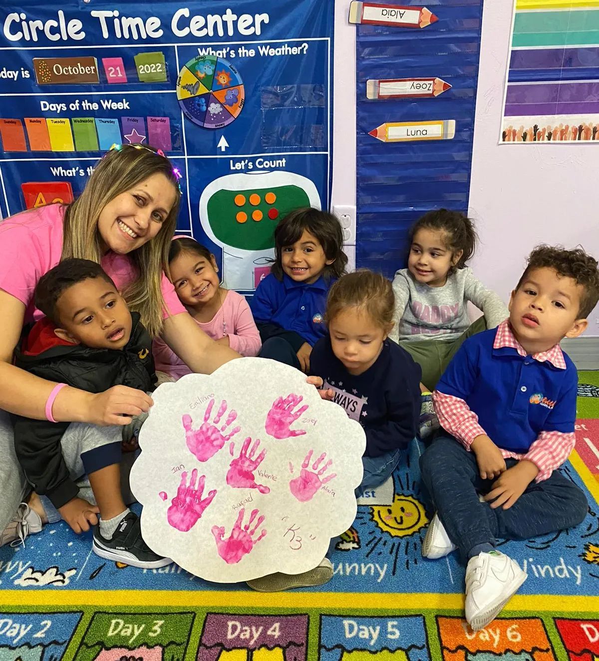 A teacher and six young children sit on a classroom rug with a handprint art project. The teacher smiles, and the children look at the camera or the art.