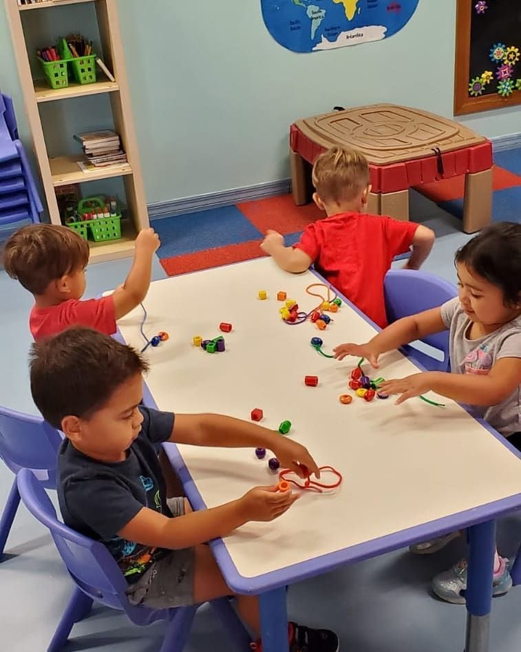 Four young children at a table stringing beads. They are in a brightly-colored classroom.