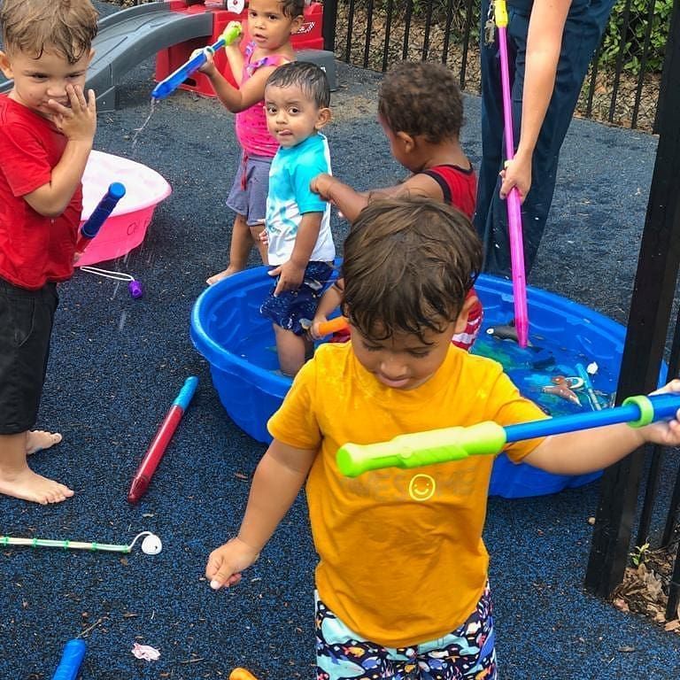 Children playing with water guns and buckets in a splash area. Several kids are wet and smiling, enjoying the water on a sunny day.