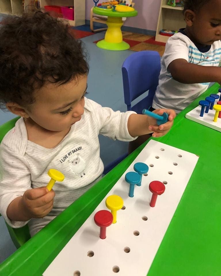 A young child in a nursery puts pegs into a board. The child is focused and holds a blue peg, with other colored pegs already in the board.