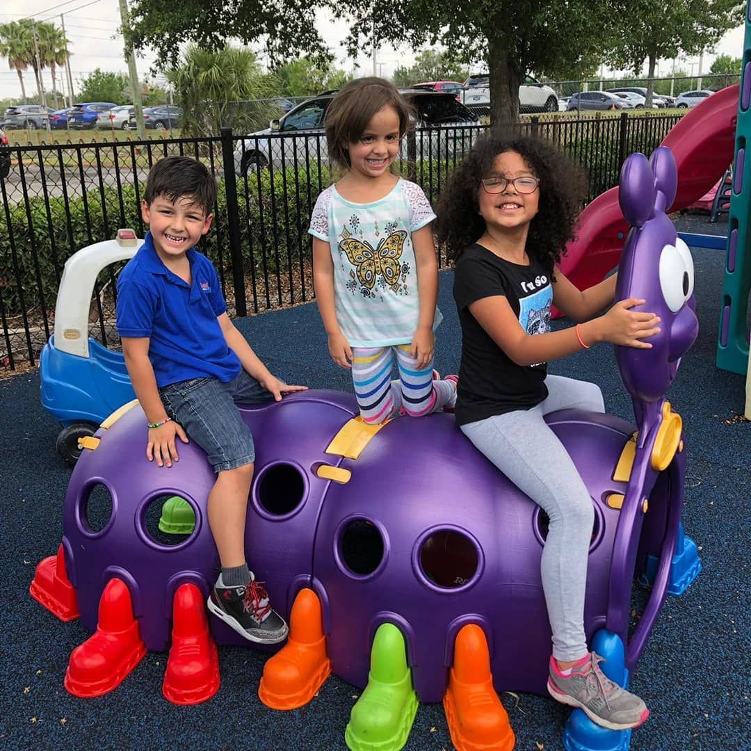 Three children smiling on a purple caterpillar playground structure. The boy wears blue, the girl wears light-colored clothing, and the other girl wears a black shirt and glasses. The playground is outdoors.