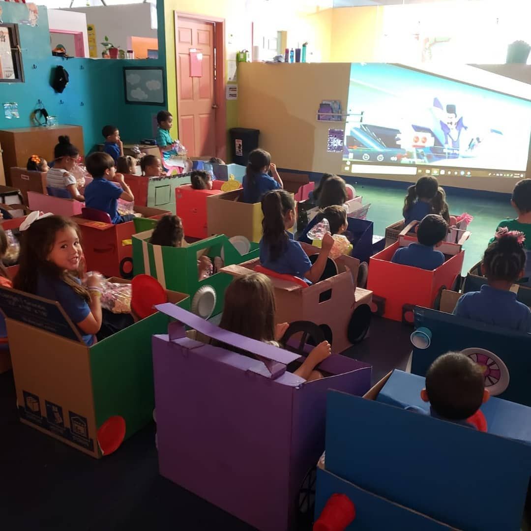 Children watch a movie from cardboard box cars in a colorful classroom. Some children smile.