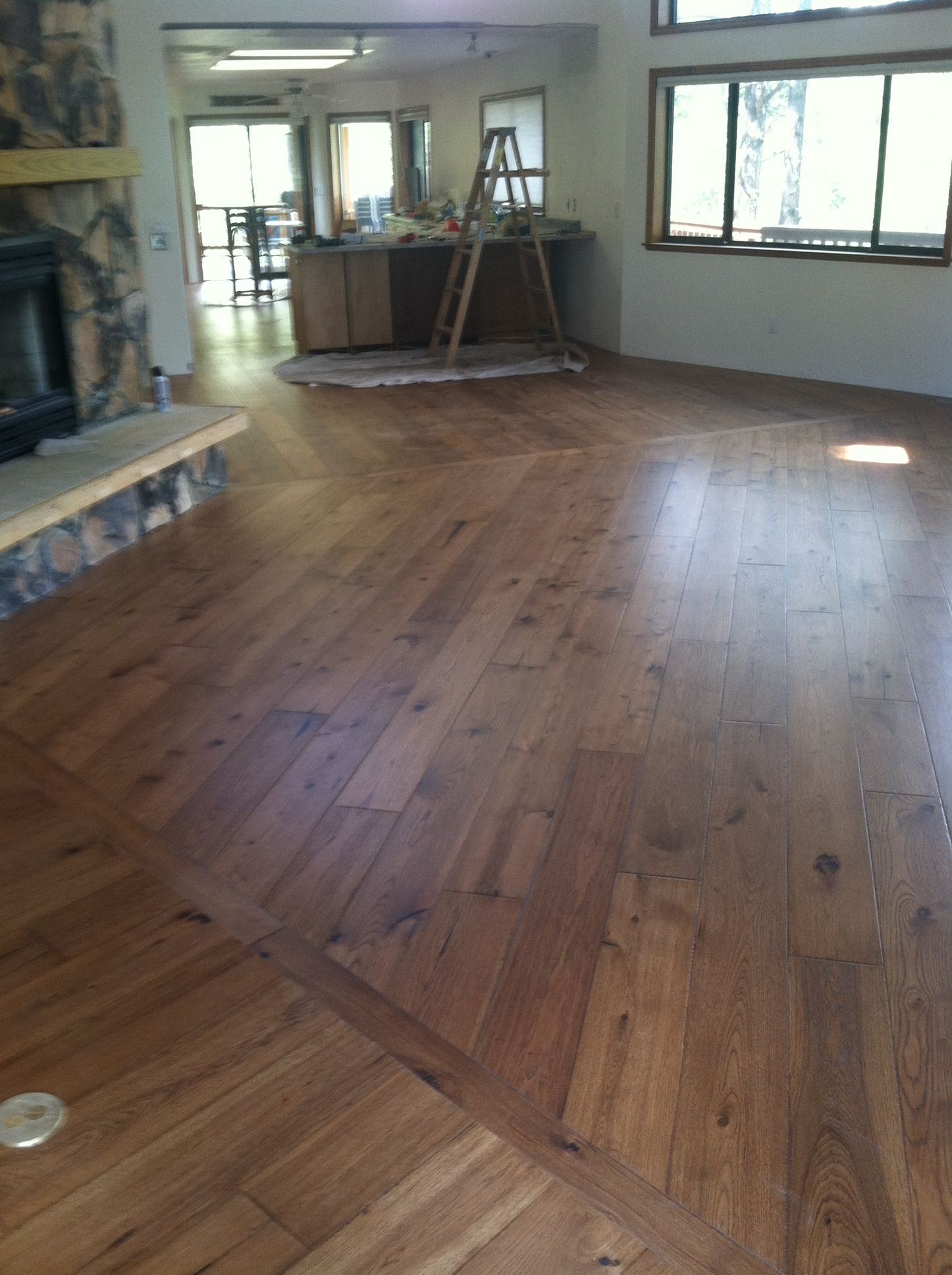 Empty living room with polished hardwood floors, large windows, and a step ladder near the back wall