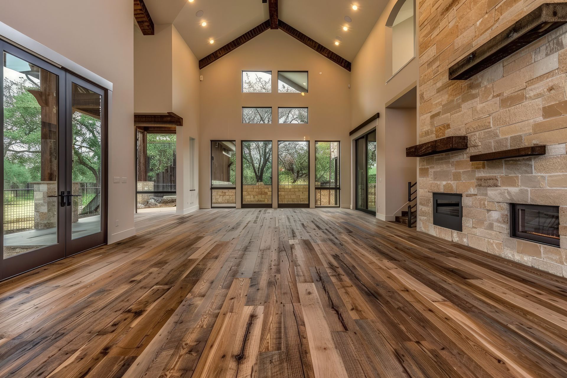 A bright, spacious room with a rustic wood floor, high vaulted ceiling, and a prominent stone fireplace wall.
