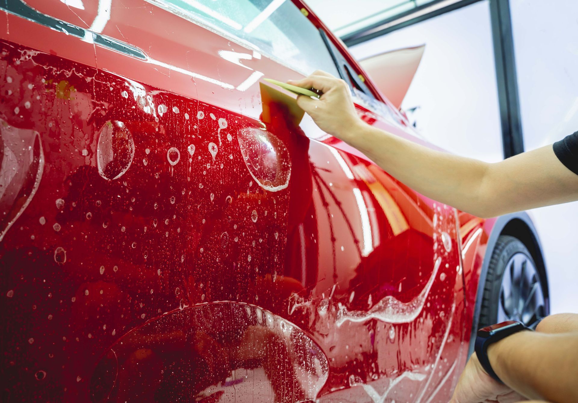 Worker applying a protective film to a red car door, smoothing it with a squeegee tool.