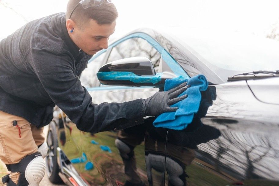 A person wearing black gloves wipes the side mirror of a black car with a blue microfiber cloth.