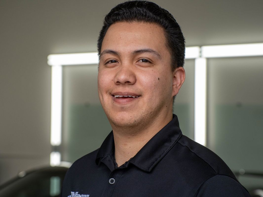 A man in a blue shirt is standing in front of a car and smiling.