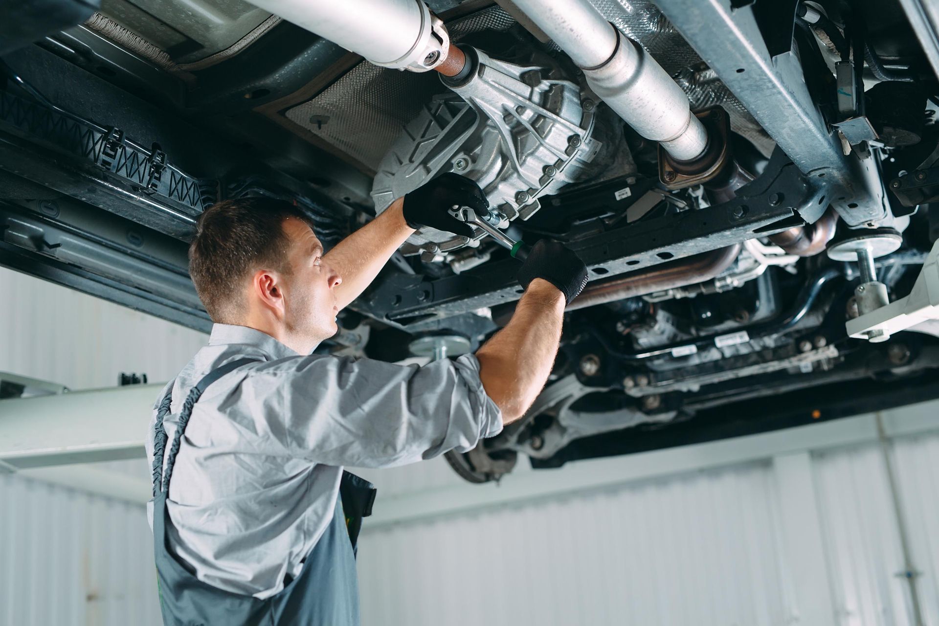 A mechanic wearing gloves and grey work clothes repairs the underside of a vehicle lifted in an auto shop.