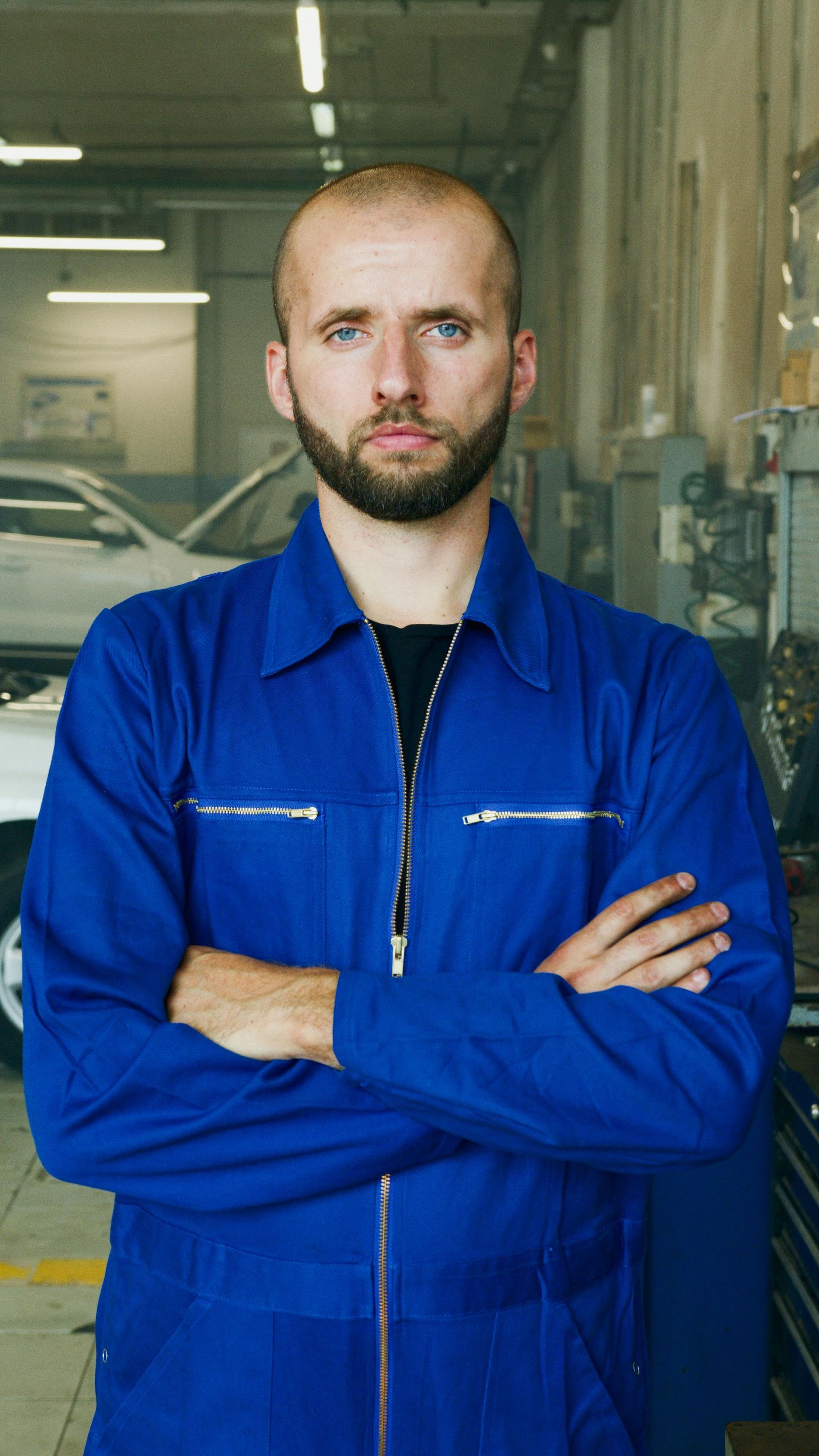 A person with a beard, wearing a blue jumpsuit, stands with arms crossed in an auto repair shop.