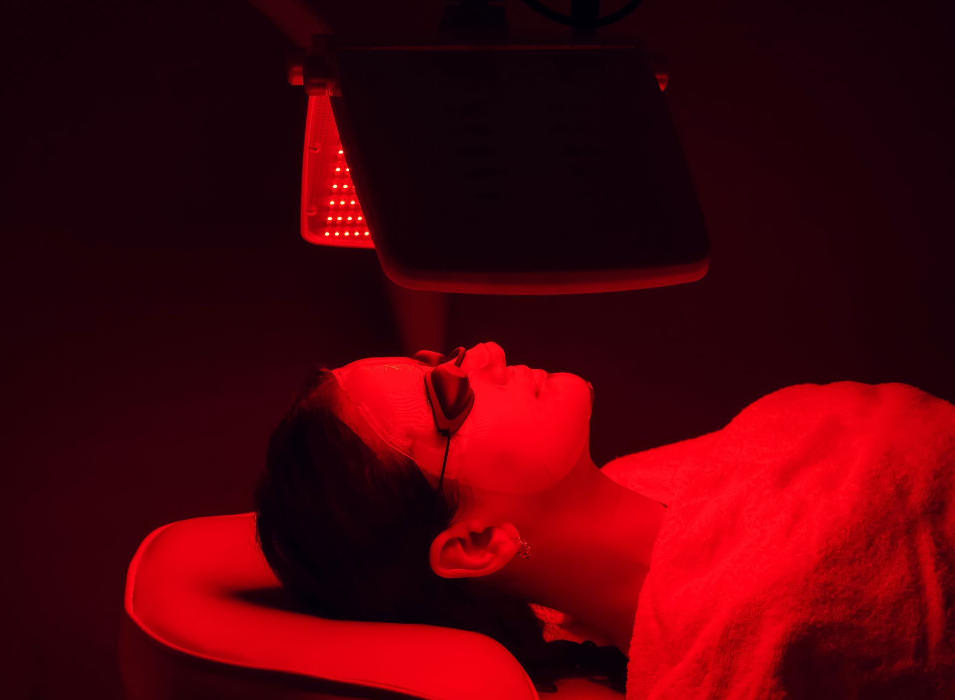 Person receiving red light therapy treatment; face covered with protective eyewear, lying on a white towel.