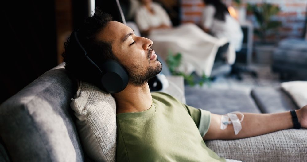 A person reclines on a couch with headphones on, resting with their eyes closed and a bandage on their inner arm.