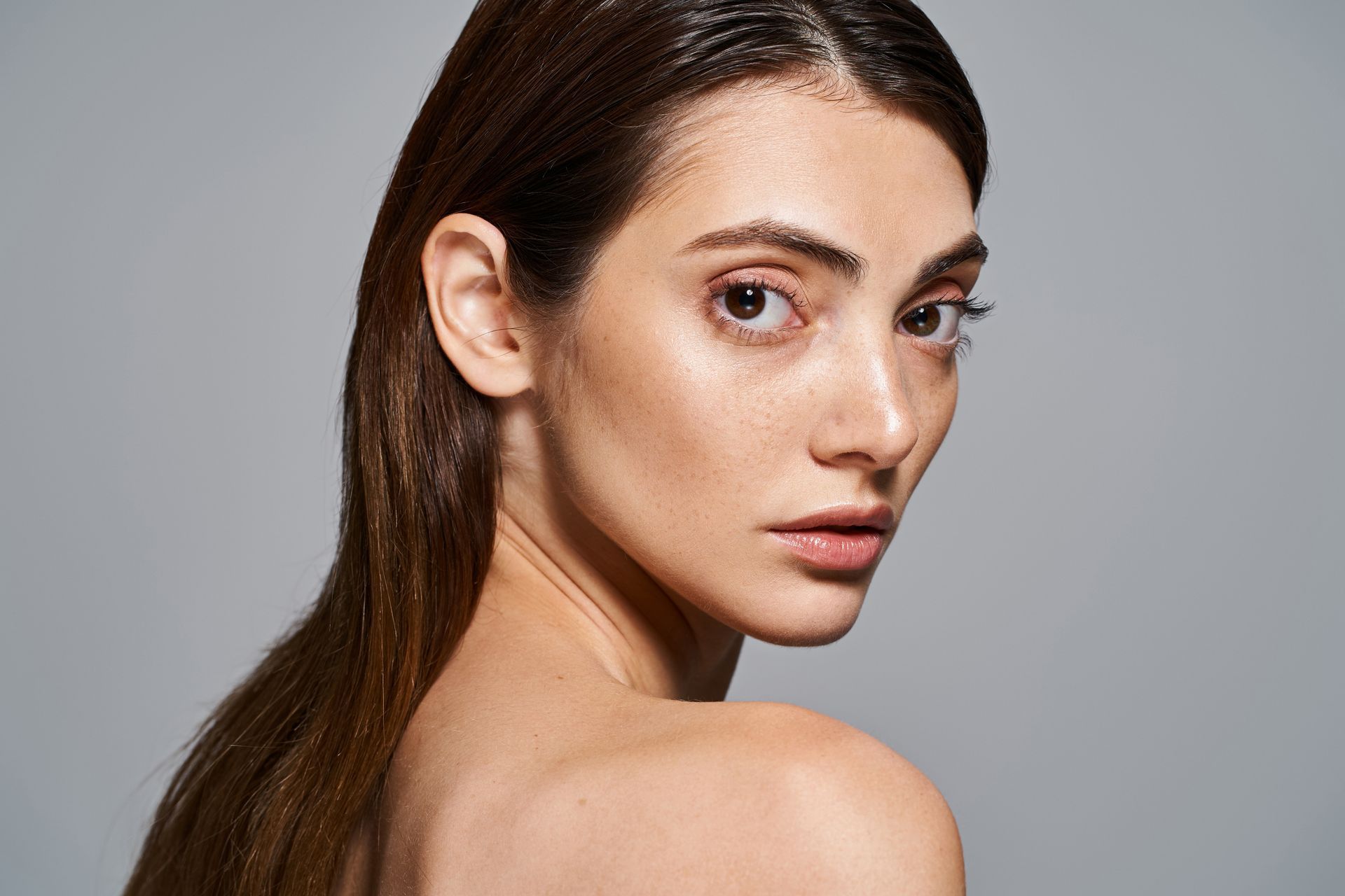 Woman with wet, slicked-back brown hair, looking over shoulder, bare shoulders, neutral skin tone.