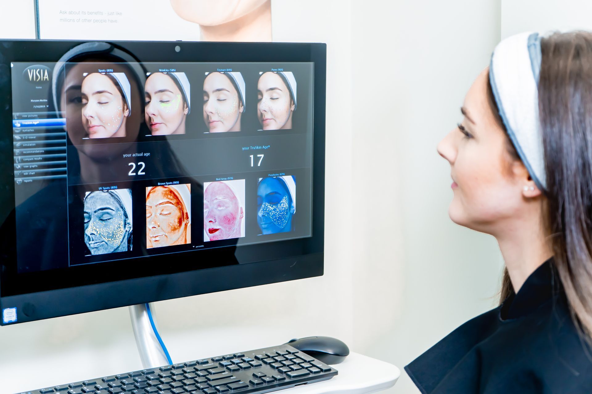 Woman using facial analysis machine; screen displays skin analysis with overlaid graphics, setting is medical office.