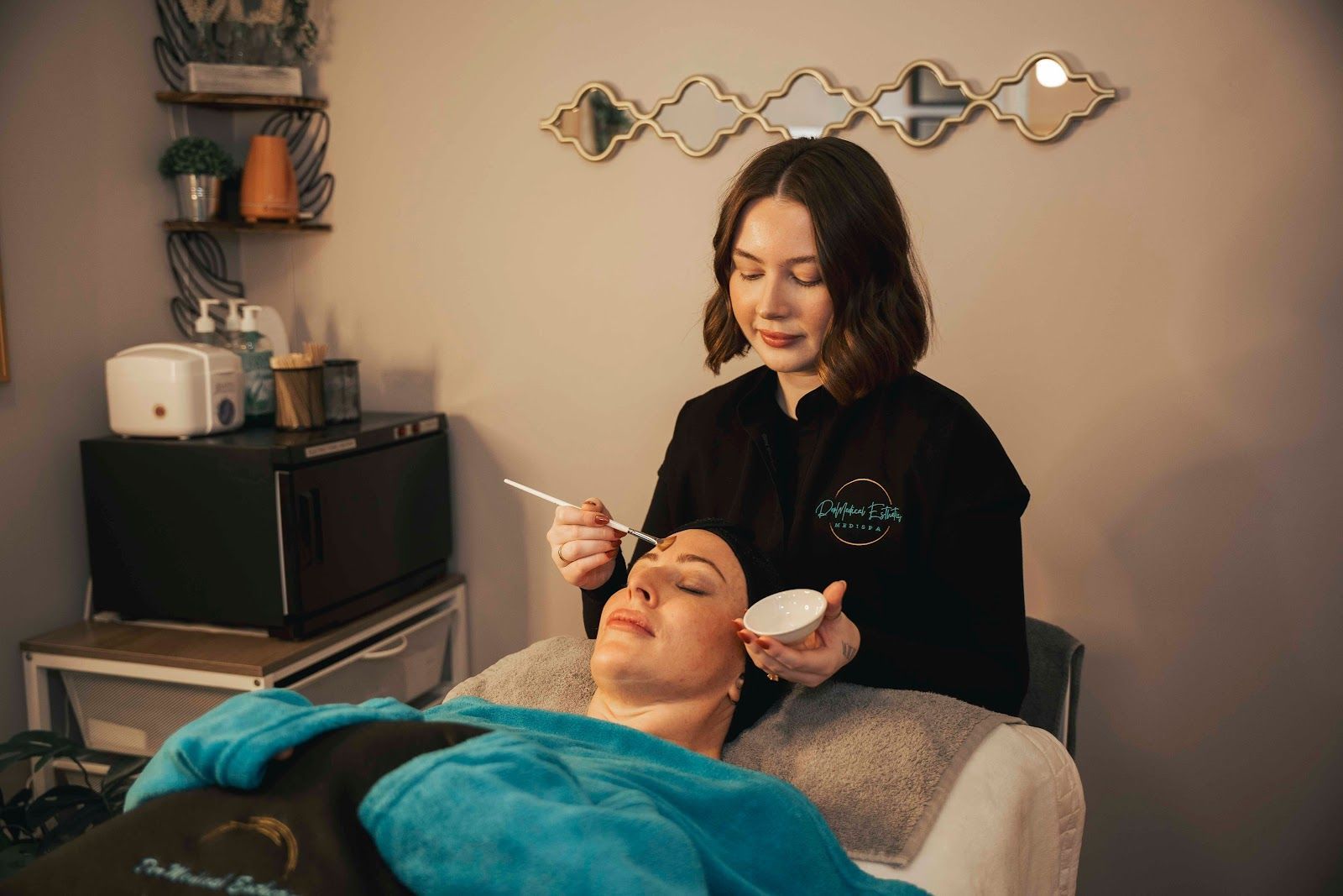 A professional applying a treatment to a client's eyebrow with a small brush and bowl in a spa treatment room.