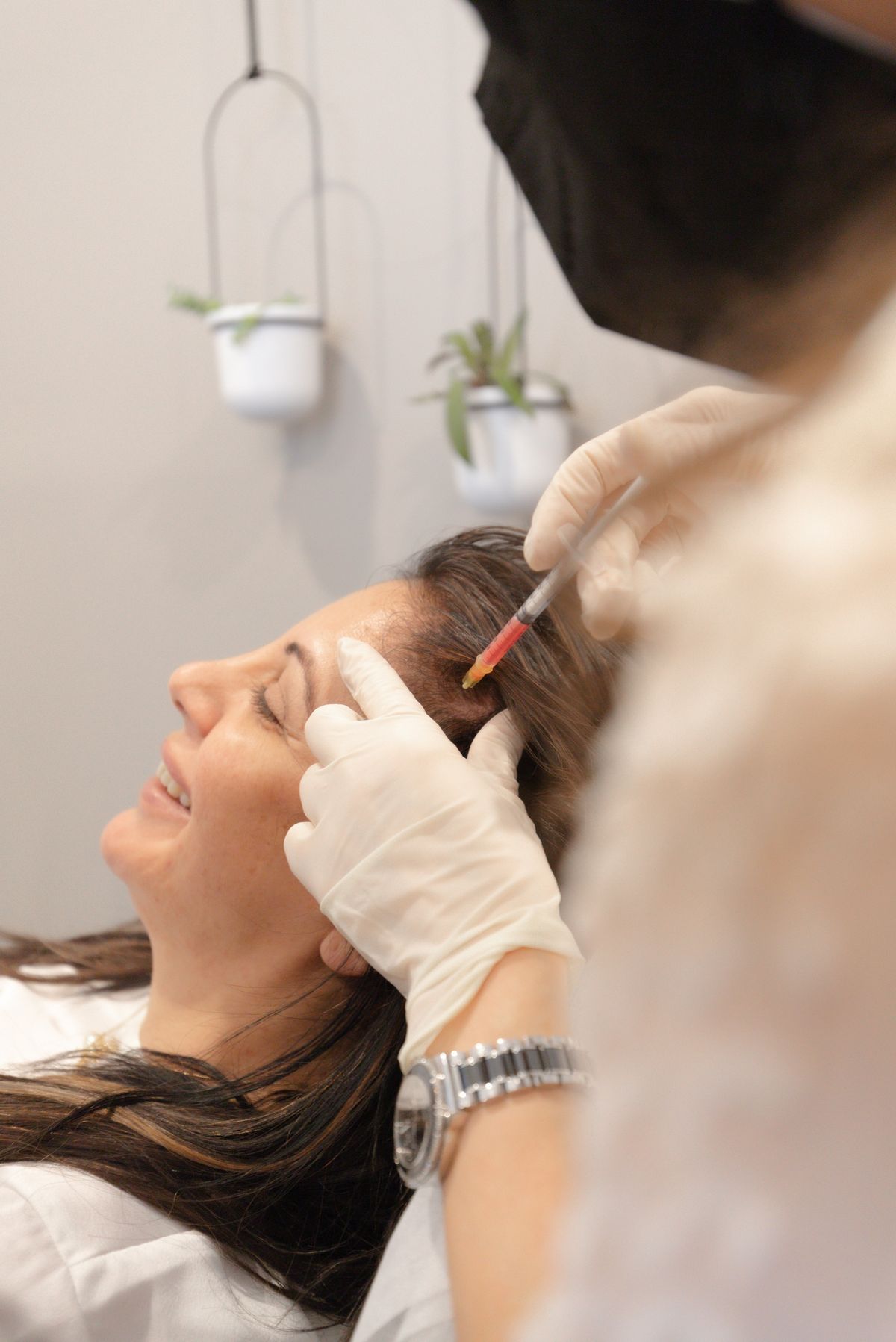 Woman receiving injection in her forehead, being performed by gloved hand. She smiles, in a clinic setting.