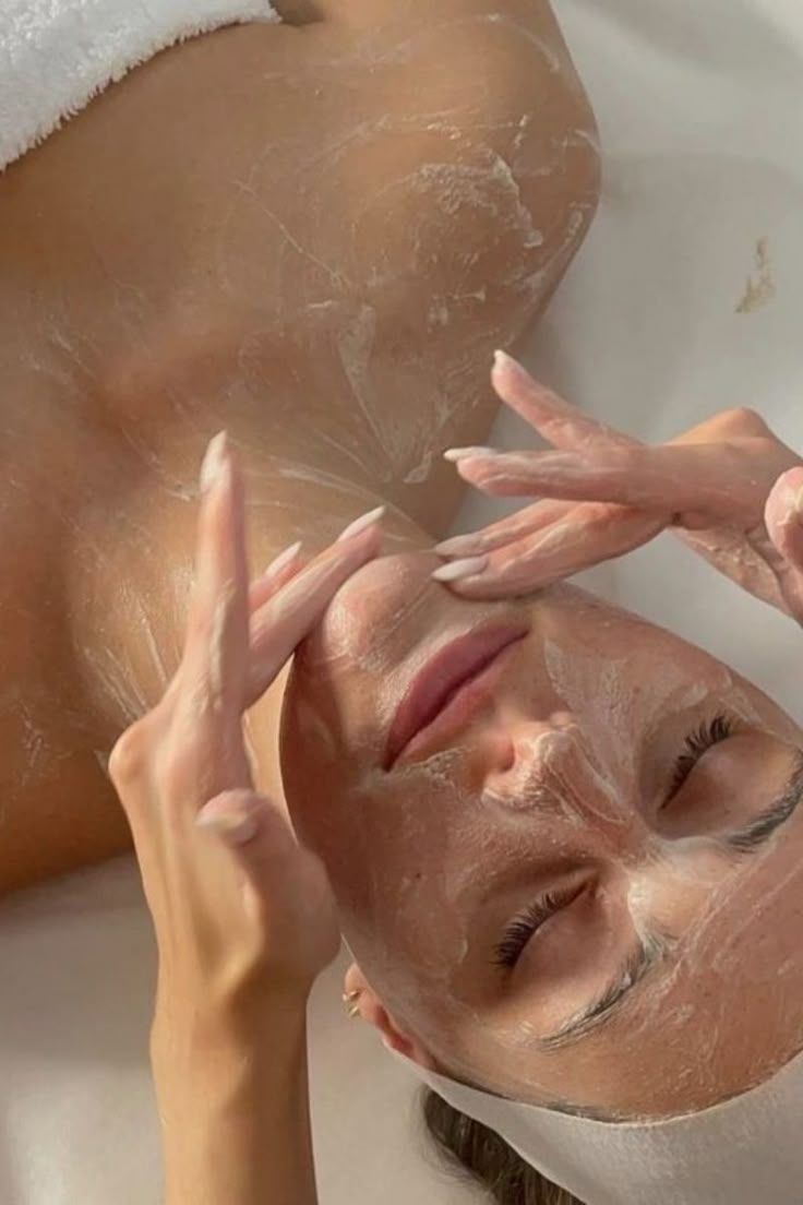 Woman receiving facial treatment, lying down with eyes closed; hands massaging face with cream.